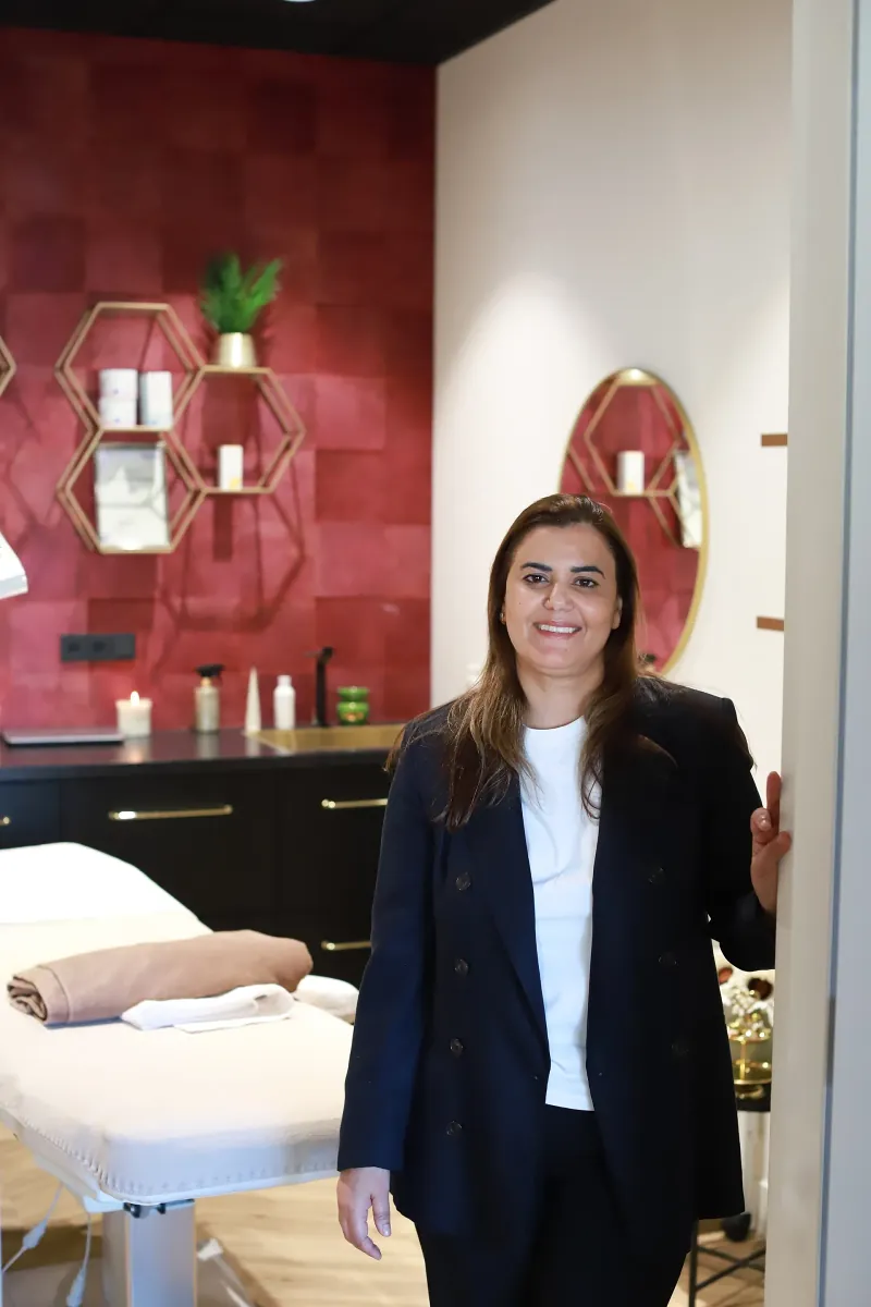 Soumaya in een donker pak in de deuropening van haar behandelkamer, haar hand op de deurpost
