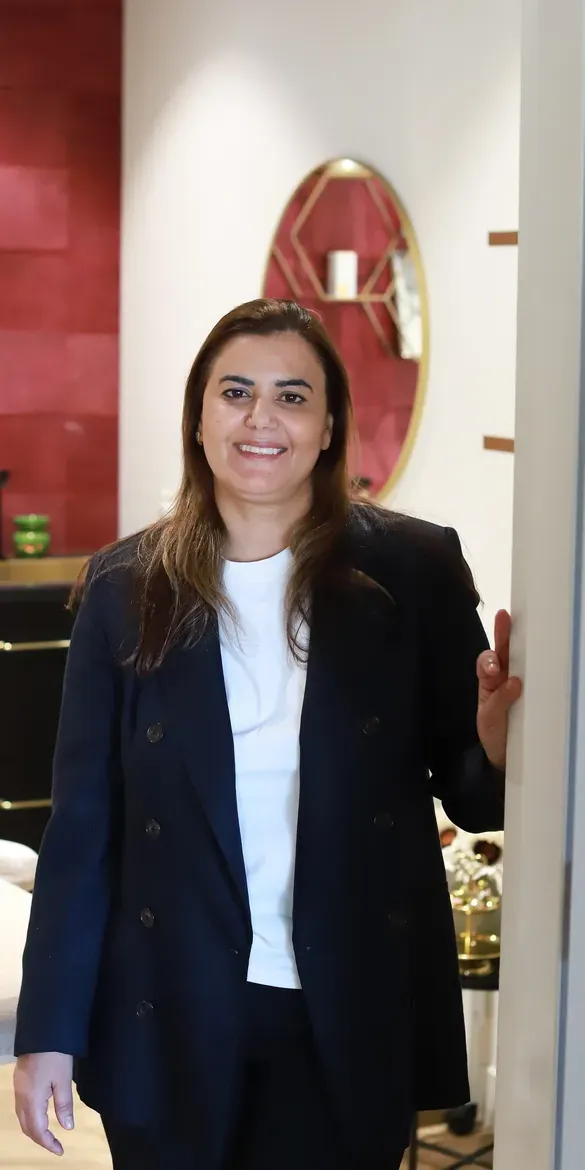Soumaya in de deuropening van haar behandelkamer, haar hand op de deurpost; achter haar de wijnrode tegelwand en het bed
