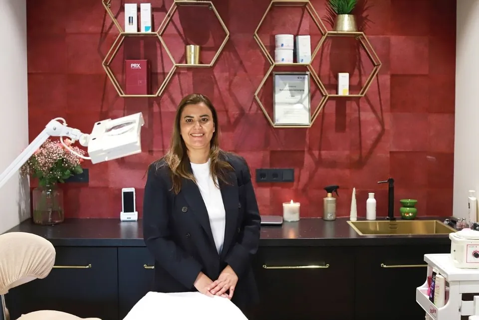 Soumaya lachend in een blazer voor de wijnrode wand en de zeshoekige planken